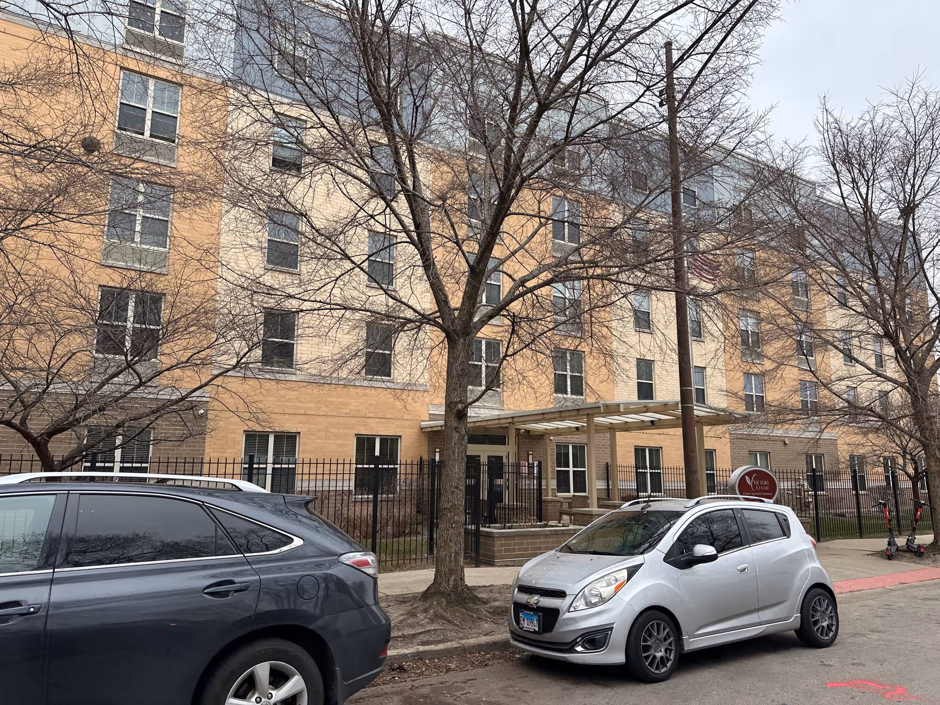 Front exterior of a multi-story brick senior living building with parked cars and leafless trees along the sidewalk.