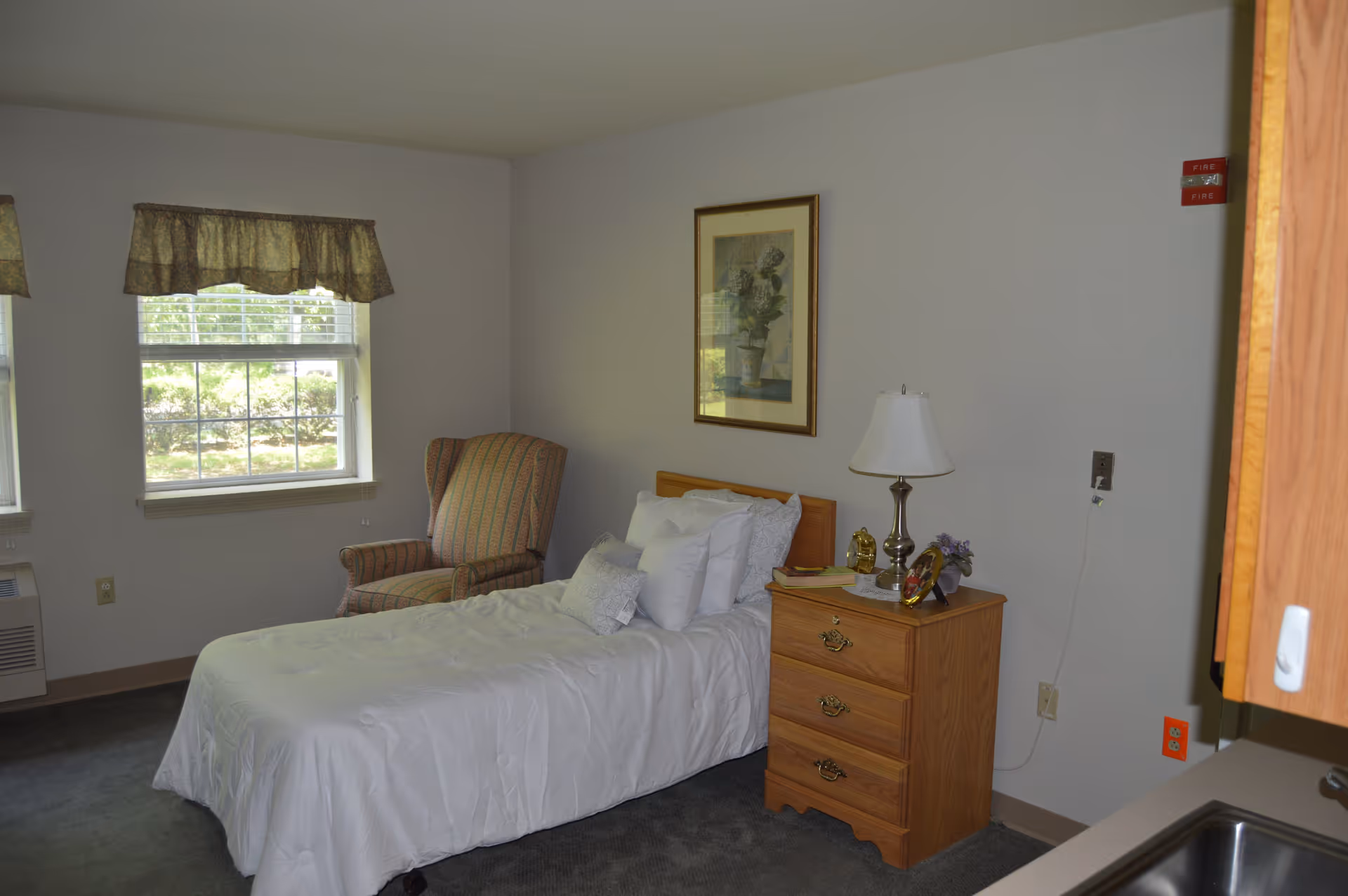 A simple bedroom with a single bed covered in white bedding, a wooden nightstand with a lamp, and a framed picture on the wall. There is a patterned armchair near two windows with valances, and part of a kitchen sink and cabinet is visible on the right side.