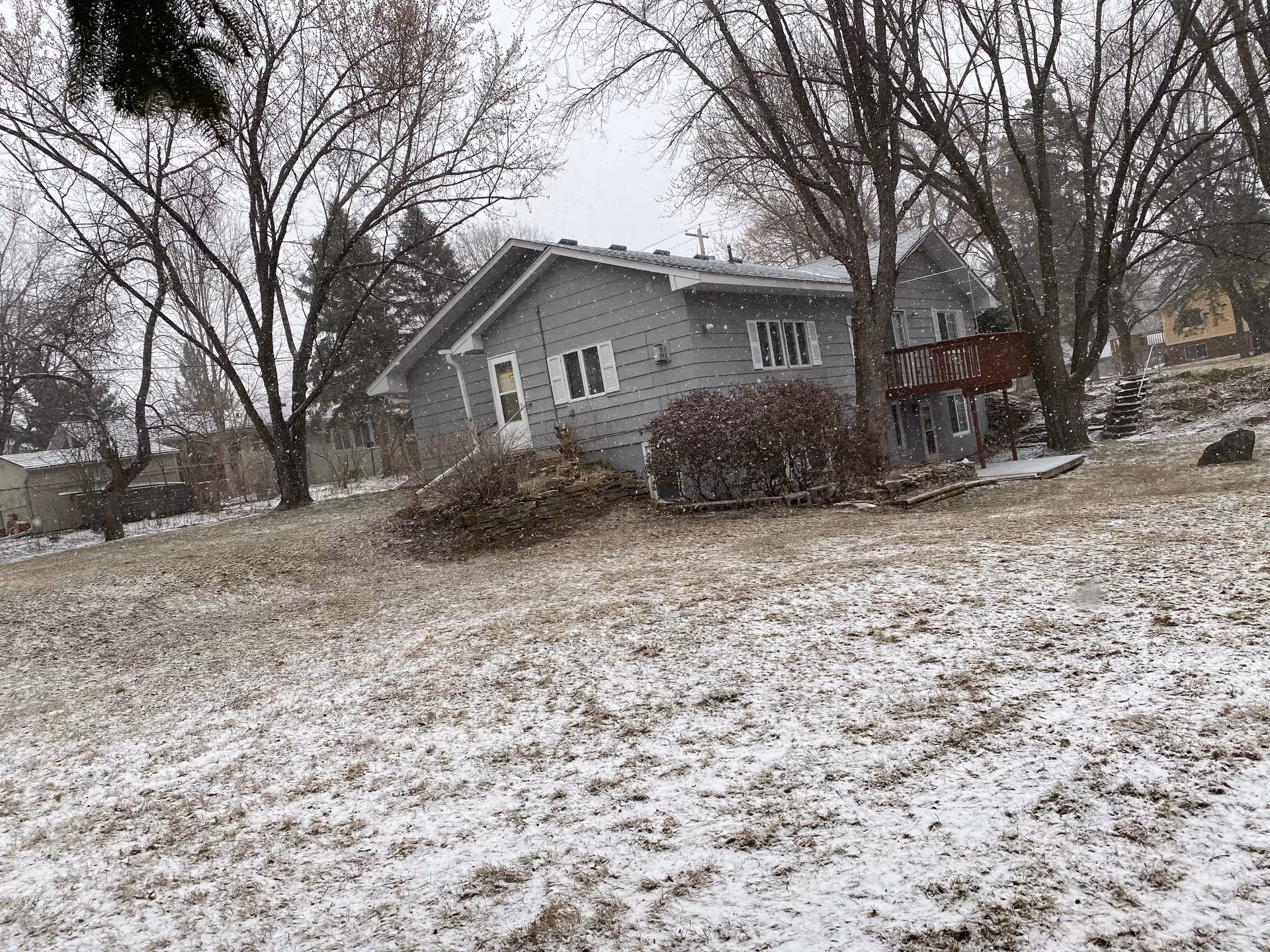 Gray single-story house with a raised wooden deck and leafless trees in a snowy yard during light snowfall.