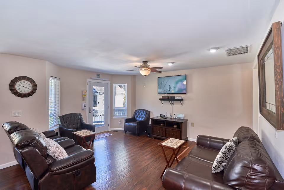 A cozy living room with dark leather sofas and armchairs arranged around two small wooden tables. A wall-mounted TV is above a wooden cabinet, and a ceiling fan with a light fixture is on the ceiling. There is a large wall clock on one wall and a framed mirror on another. The room has hardwood floors and windows letting in natural light.