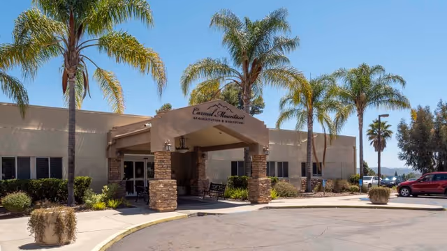 Exterior view of Carmel Mountain Rehabilitation And Healthcare Center building entrance with palm trees, a covered drop-off area supported by stone pillars, and a parking lot with a red vehicle visible.