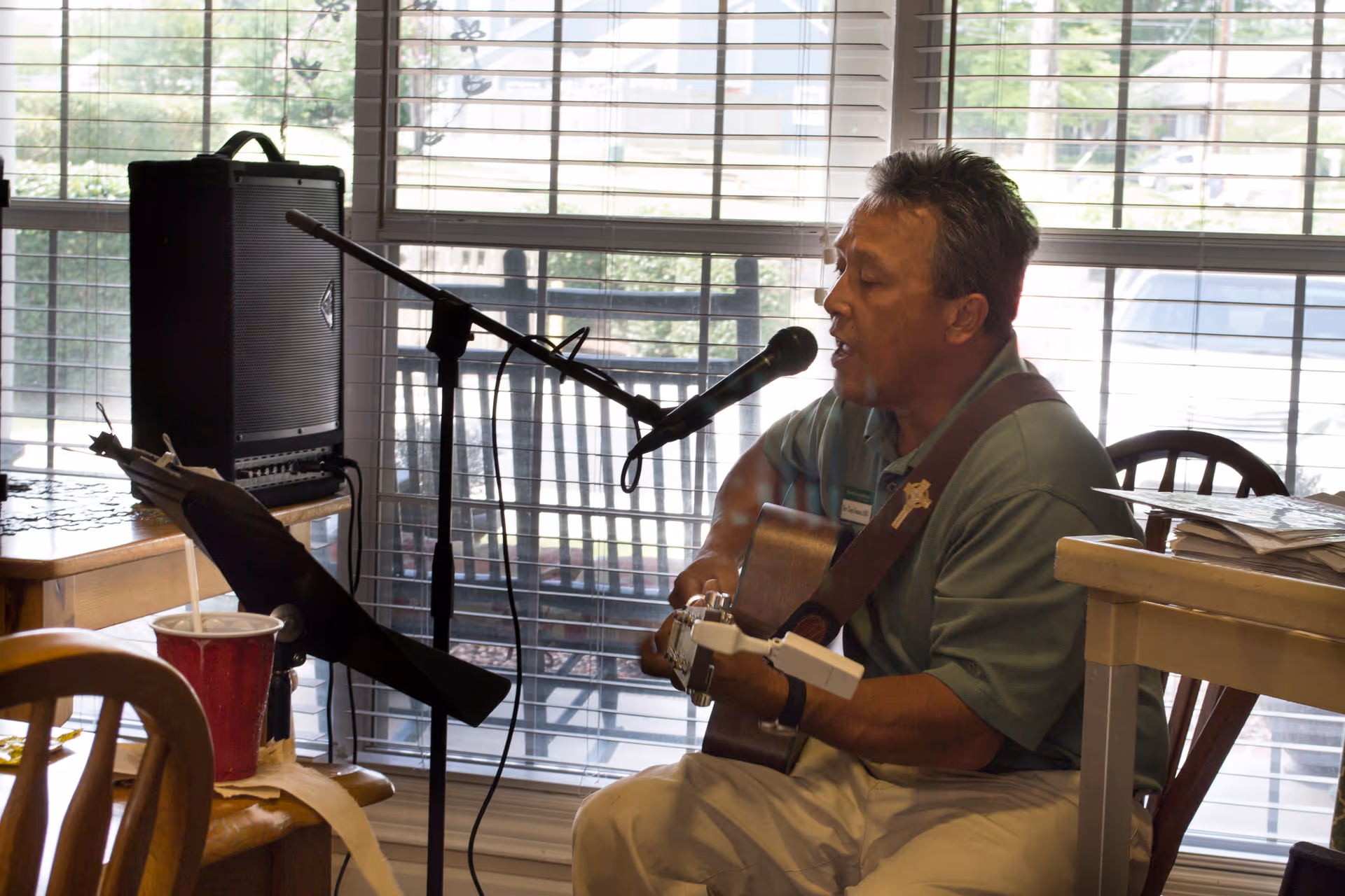 A man playing an acoustic guitar and singing into a microphone in a room with large windows covered by horizontal blinds. There is a speaker, a music stand with sheet music, and a red cup on a nearby table.