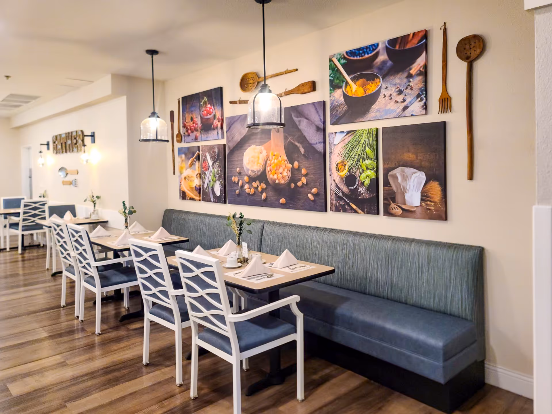 Dining area with a long blue cushioned bench along the wall and white chairs with blue seats around tables set with napkins, plates, and utensils. The wall above the bench is decorated with framed pictures of food ingredients and wooden kitchen utensils. Pendant lights hang from the ceiling, illuminating the space.