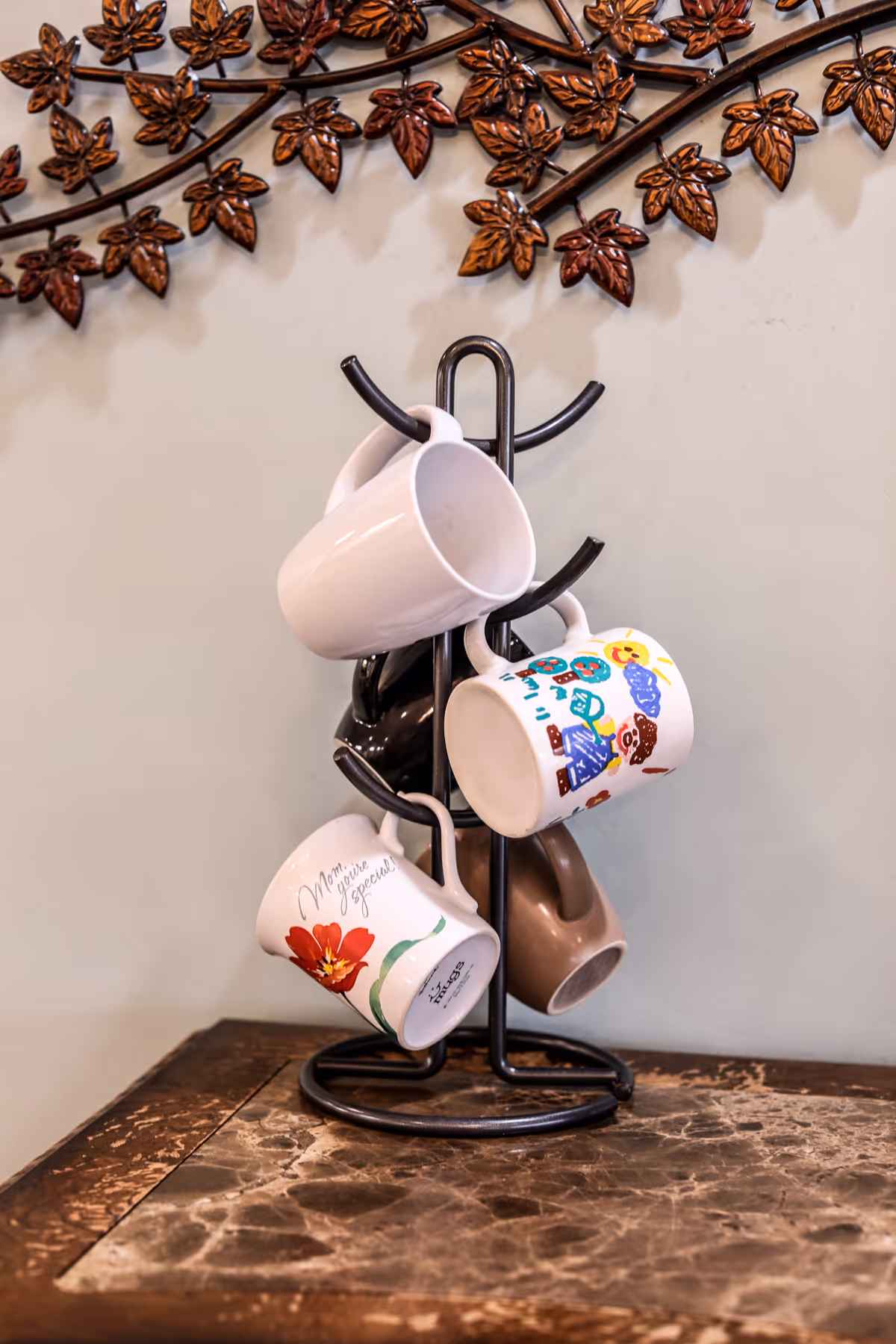 A mug tree holding several colorful and white ceramic mugs on a wooden table beneath decorative metal leaf wall art.