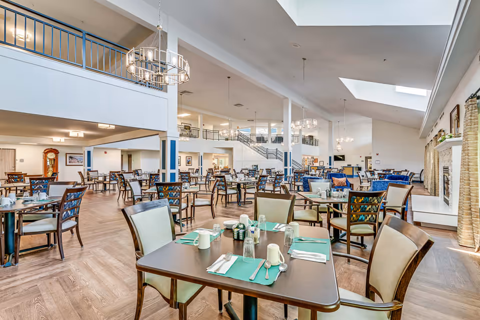 A spacious and well-lit dining room in a senior living facility with multiple tables and chairs arranged neatly. The tables are set with placemats, utensils, cups, and glasses. The room features high ceilings with modern chandeliers, large windows with curtains, and a fireplace on the right side. There is a mezzanine level with railings and a staircase leading up to it.