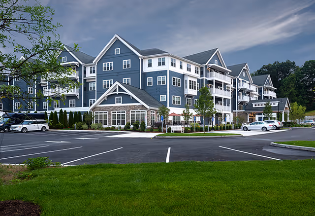Exterior view of a large, multi-story senior living facility building with blue siding and white trim, surrounded by a parking lot with several cars and green landscaped areas under a cloudy sky.