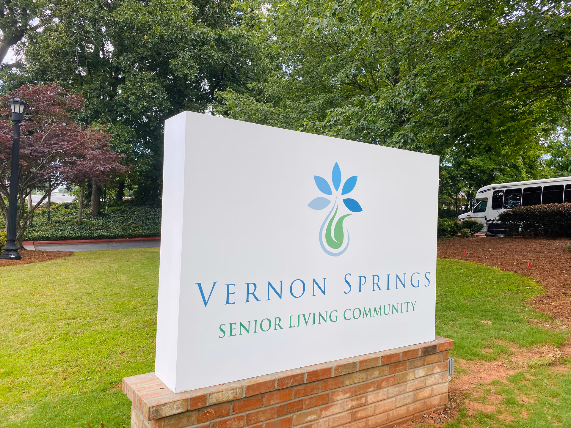A large white sign with a blue and green floral logo and the text 'Vernon Springs Senior Living Community' stands on a brick base in a grassy area surrounded by trees and bushes. A white shuttle bus is partially visible in the background.
