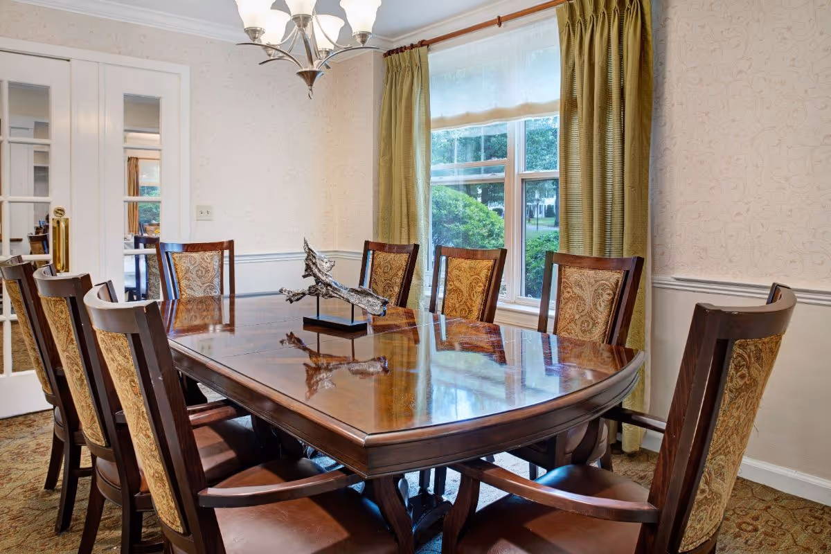 Wooden dining room table with eight upholstered chairs, a decorative centerpiece, chandelier, and a window with green curtains.