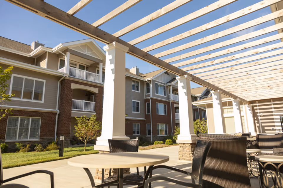 Outdoor patio area with round tables and wicker chairs under a wooden pergola, adjacent to a multi-story residential building with balconies and windows, on a sunny day.