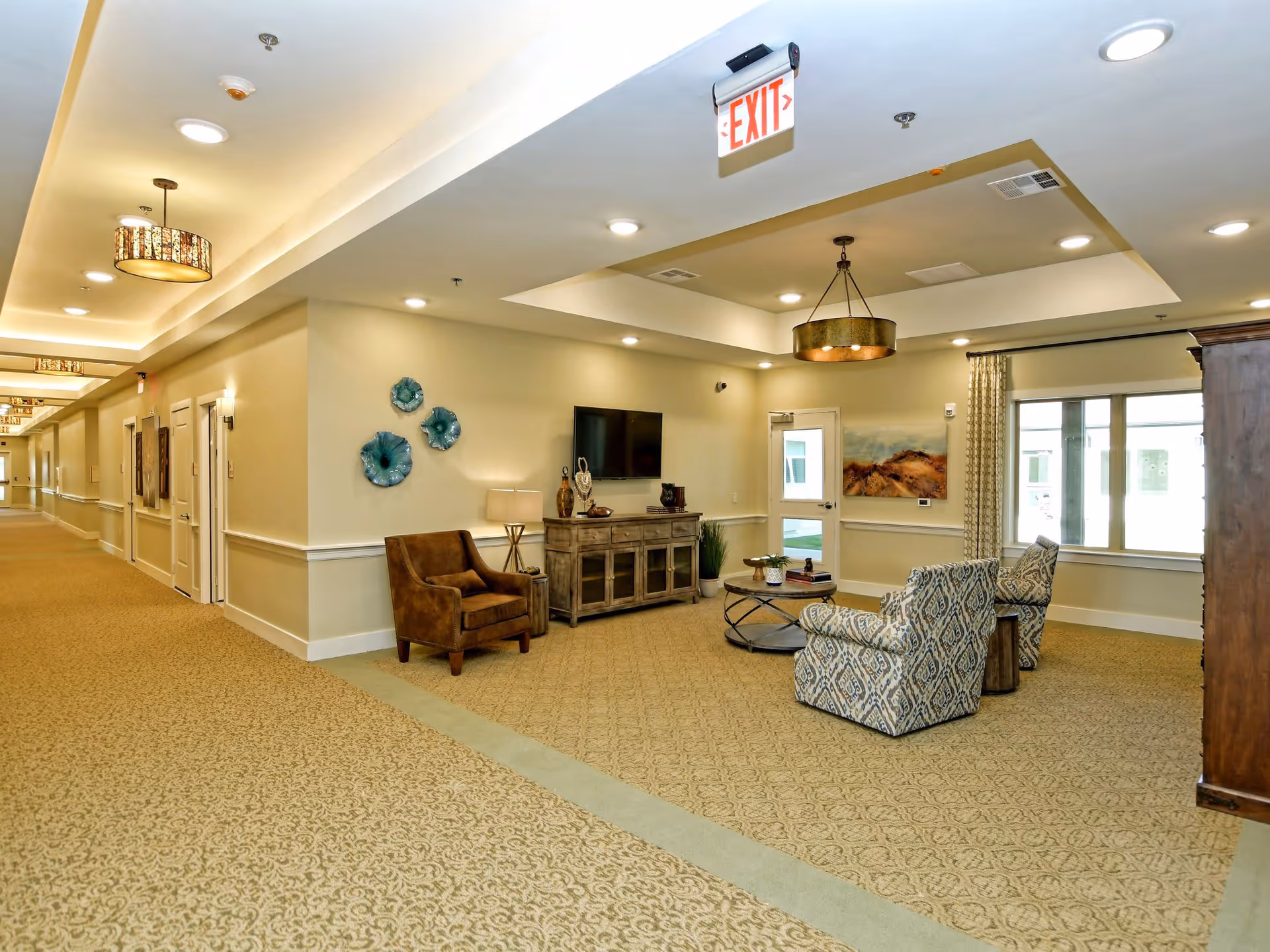 A well-lit senior living facility common area with beige patterned carpet and cream-colored walls. The space includes a seating area with two patterned armchairs, a brown armchair, a round coffee table, a wooden cabinet with a flat-screen TV mounted above it, and decorative wall art. Ceiling lights and a hanging light fixture illuminate the area. A hallway extends to the left with multiple doors and wall decorations. An exit sign is visible on the ceiling.