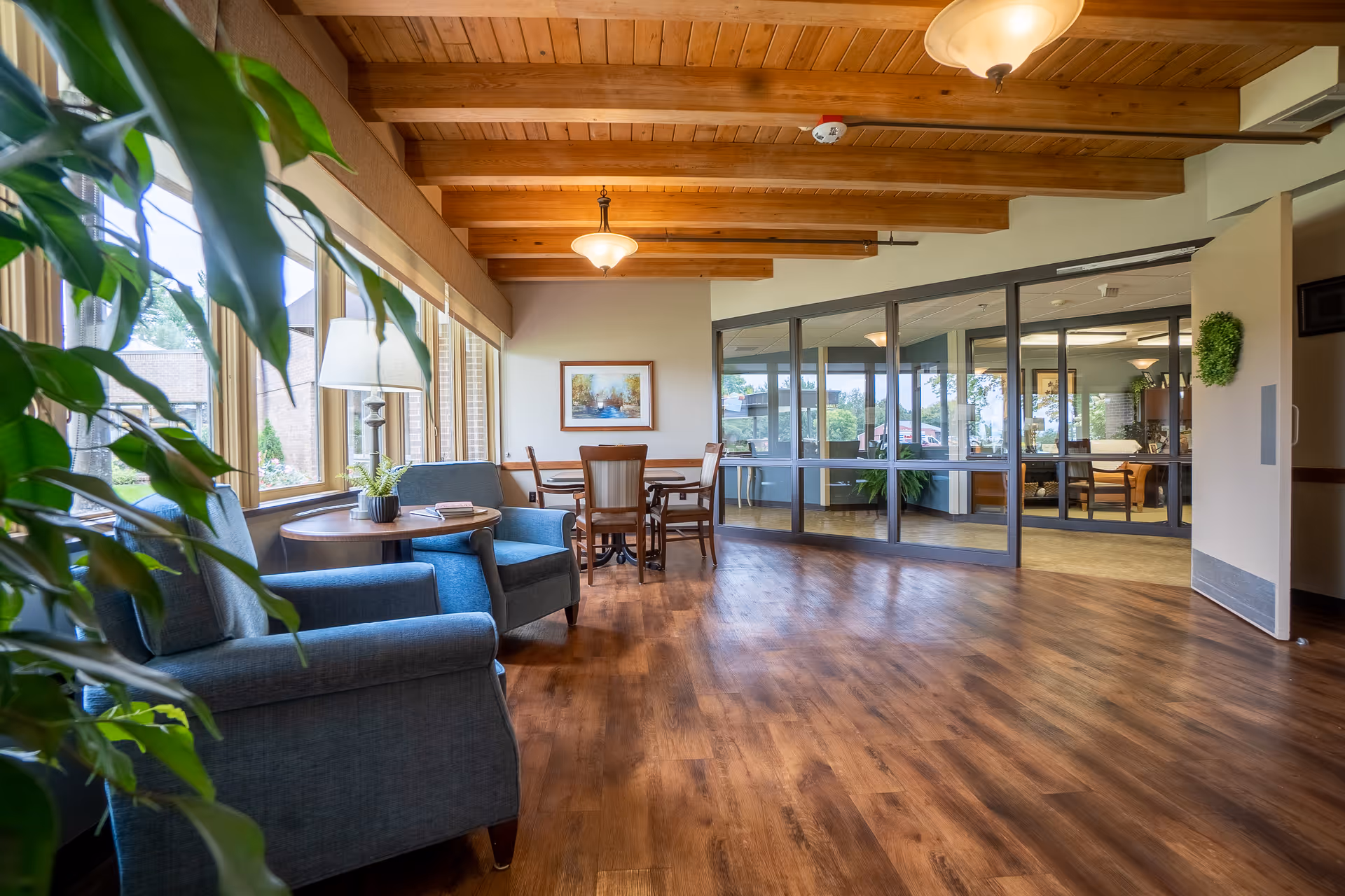 Bright communal seating area with armchairs, a small table, wooden-beamed ceiling, and large windows in a senior living facility.