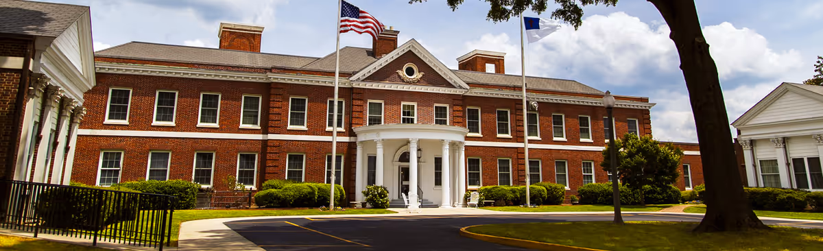 Front exterior view of Shepherd's Care Assisted Living Center, a large two-story brick building with white columns at the entrance, two flagpoles with the American flag and another flag, surrounded by green bushes and trees under a partly cloudy sky.