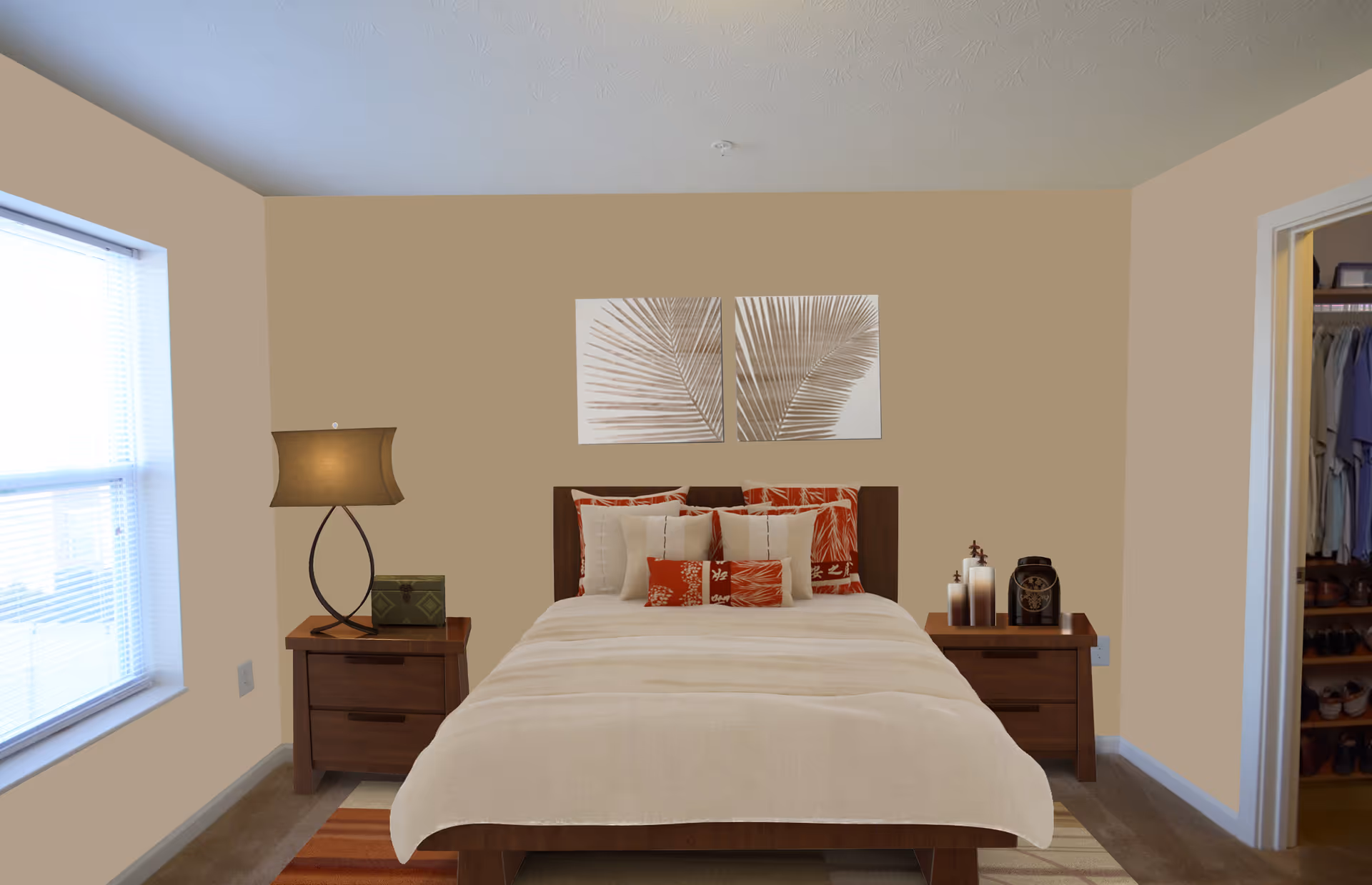 A neatly made bed with beige and red patterned pillows and a beige comforter in a bedroom. There are two wooden nightstands on either side of the bed, one with a lamp and a small box, the other with decorative candles and a clock. Above the bed are two framed artworks featuring palm leaves. A window with blinds is on the left wall, and an open closet with hanging clothes and shoes is visible on the right.