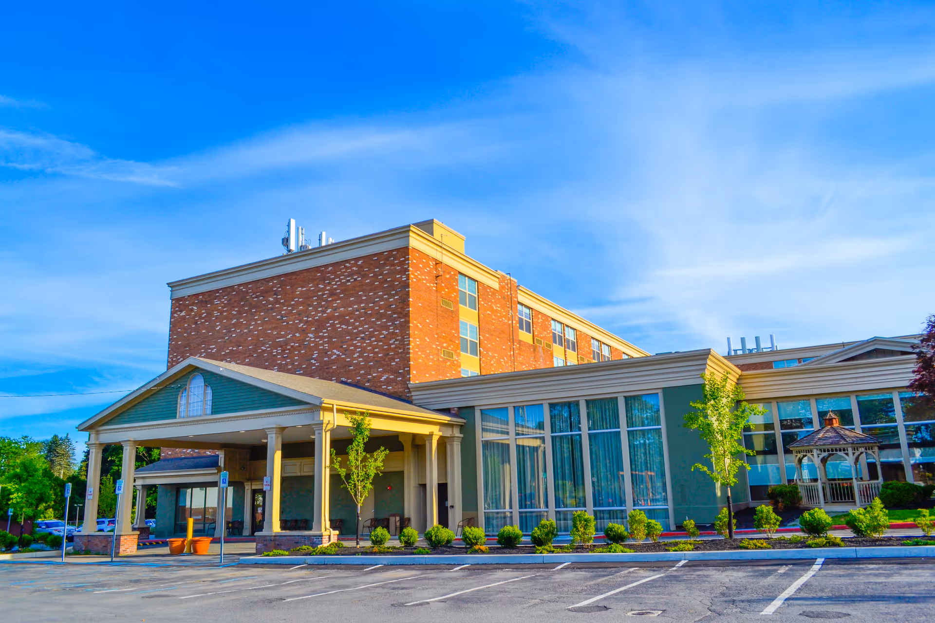 Exterior view of a senior living facility building with a brick upper section and large windows on the lower section. The entrance has a covered porch with columns, and there is a small gazebo surrounded by greenery on the right side. The sky is clear and blue.