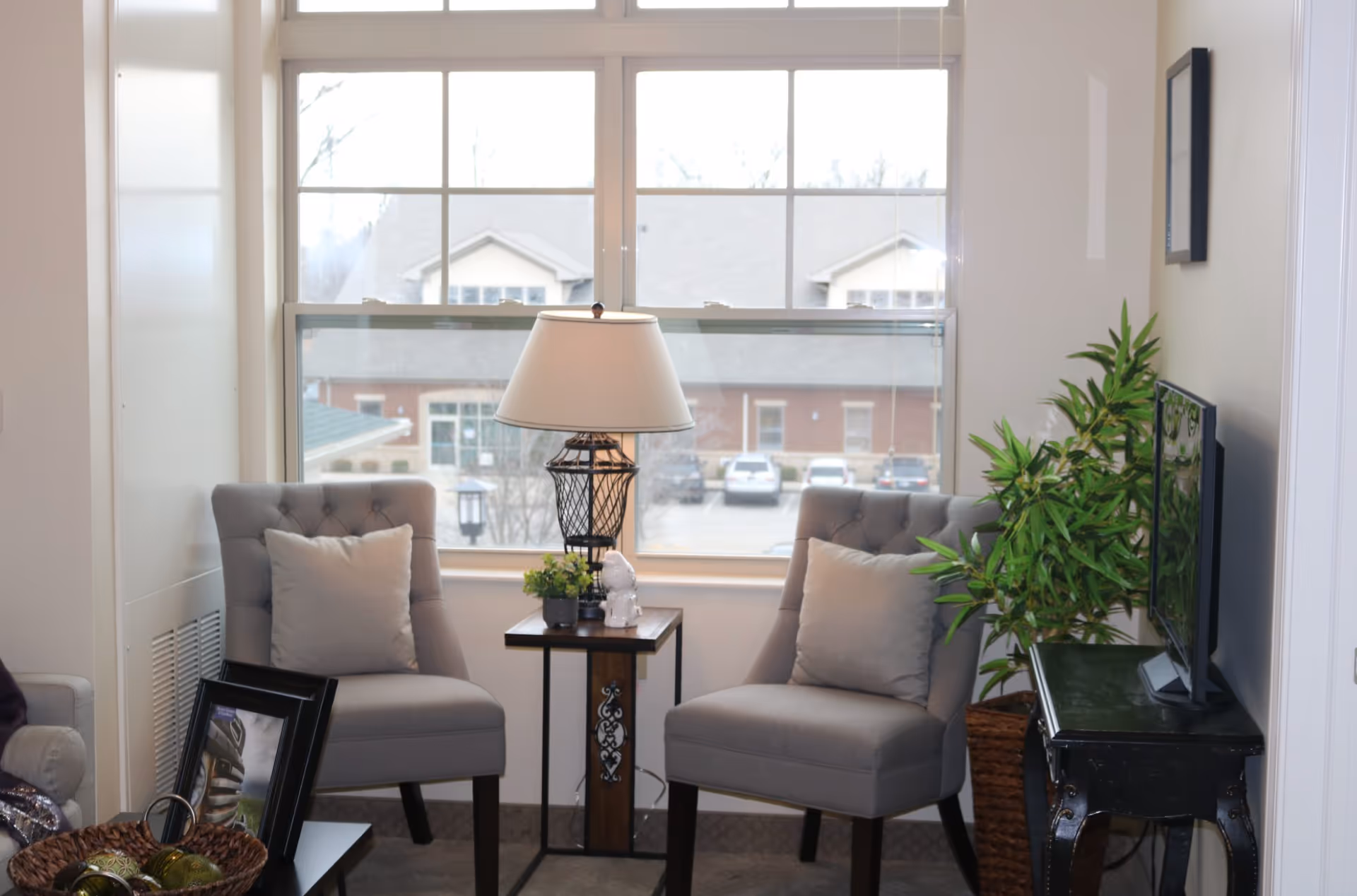 A cozy sitting area with two gray upholstered chairs with light-colored pillows, a small wooden side table with a decorative lamp and small plants, a flat-screen TV on a black table, and a large window showing a view of buildings and parked cars outside.
