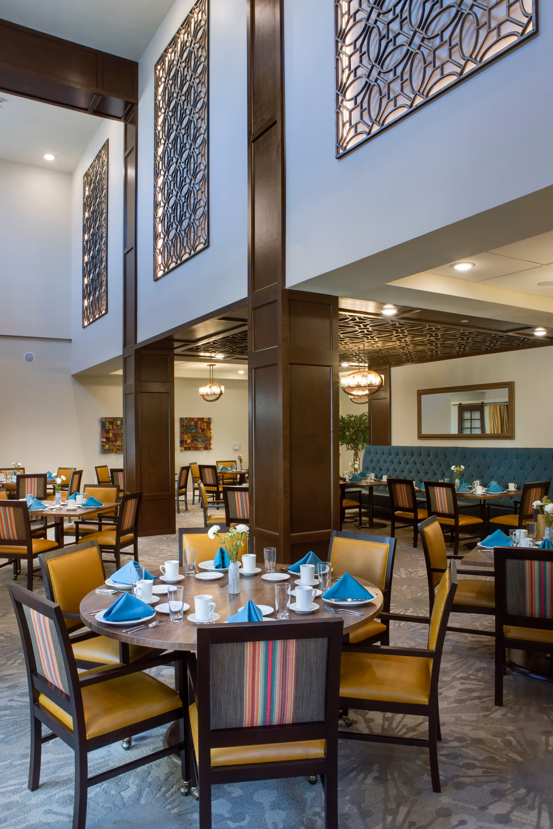 Bright, upscale dining room with round tables set with blue napkins and place settings, upholstered chairs, decorative wood columns and ornate wall panels.