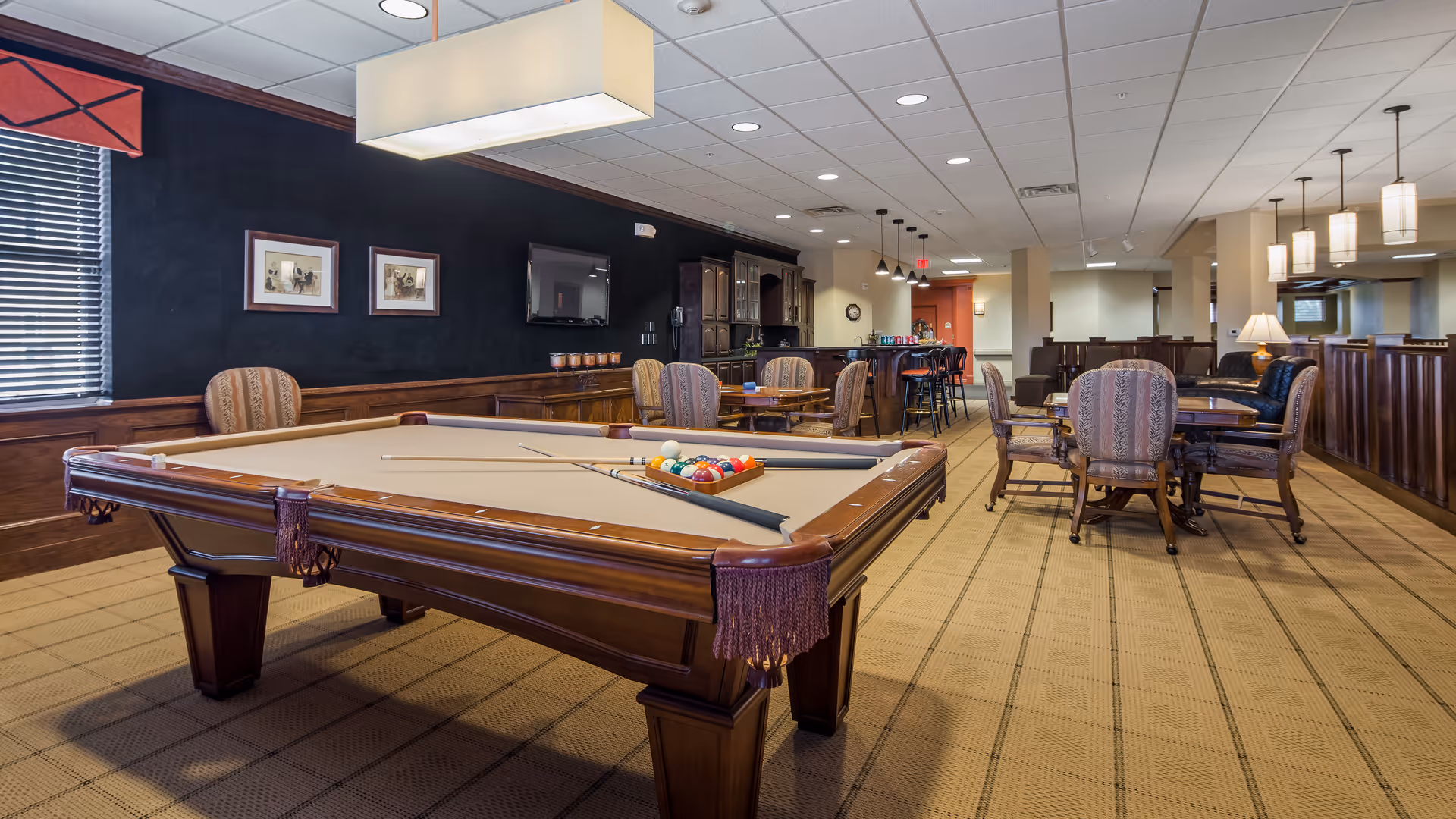 Well-lit lounge with a pool table in the foreground, upholstered chairs and tables, and a bar area in the background.