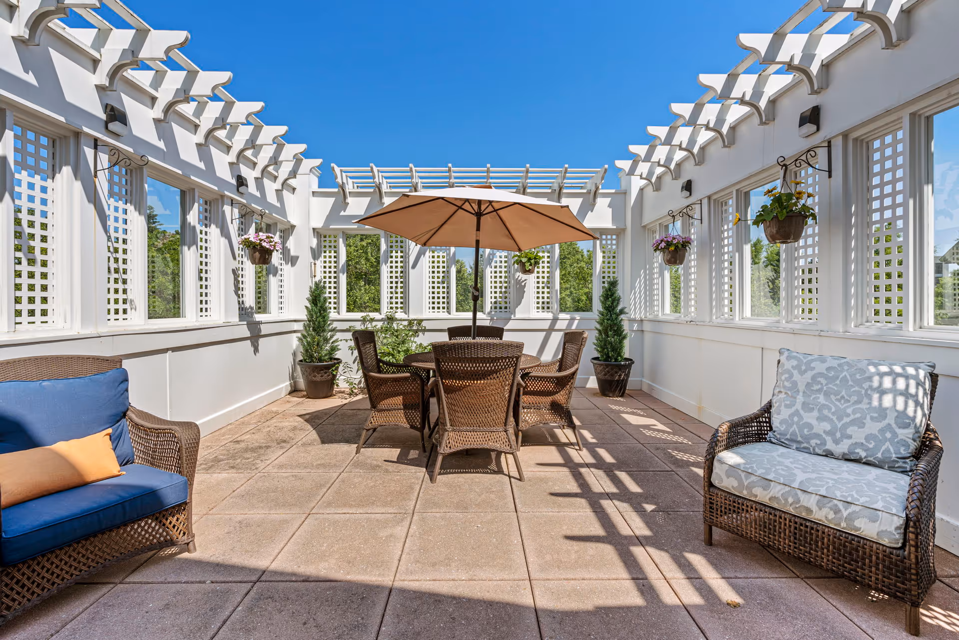Outdoor patio area with a round table and six wicker chairs under a large beige umbrella. The patio is enclosed with white lattice walls and hanging flower pots. There are two cushioned wicker armchairs, one with blue cushions and the other with light patterned cushions. The sky is clear and blue.