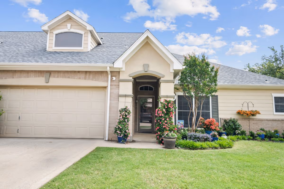 Front entrance of a single-story house with a two-car garage, landscaped lawn, and potted flowering plants.