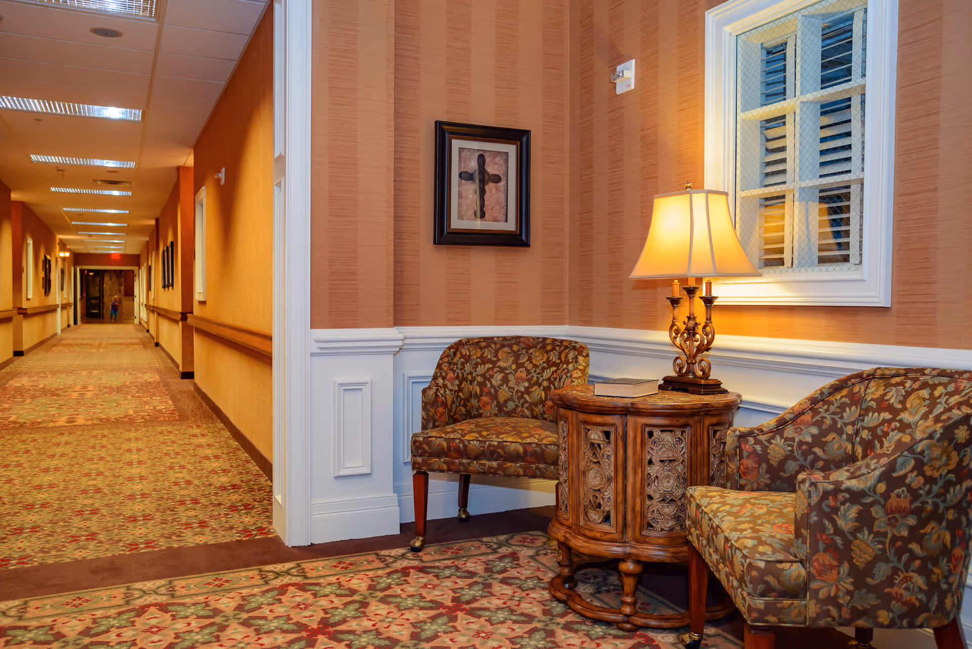 A cozy corner in a hallway of a senior living facility with two floral upholstered chairs and a carved wooden side table with a lamp and a book on it. The walls have warm-toned striped wallpaper with white wainscoting, and a framed cross artwork hangs above the chairs. The hallway extends into the distance with patterned carpet and ceiling lights.