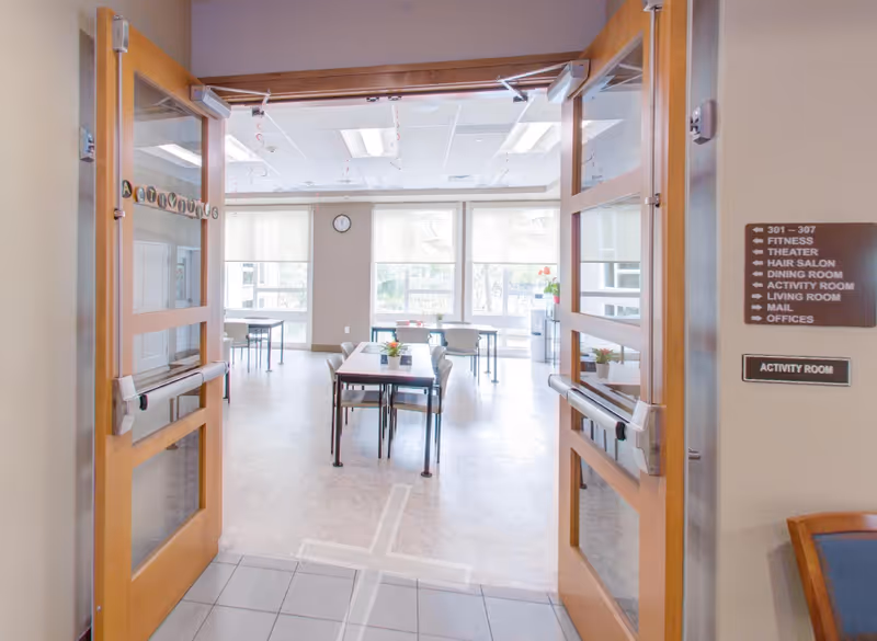 Open double wooden doors leading into a bright activity room with tables and chairs arranged neatly. Large windows at the far end let in natural light, and a clock is mounted on the wall above the windows. A sign on the right wall lists directions to various facility areas including fitness, theater, hair salon, dining room, activity room, living room, mail, and offices.