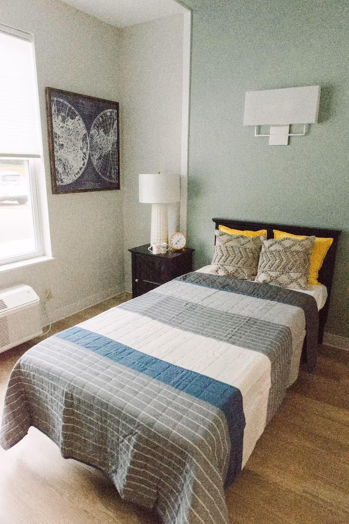 A single bed with striped bedding and decorative pillows next to a nightstand with a lamp and clock, plus wall art and a window in a bright bedroom.