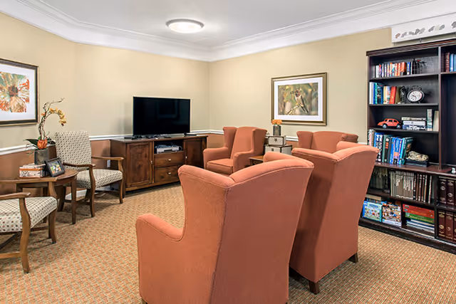 A cozy common living room with four coral upholstered armchairs facing a TV, a bookshelf, side chairs, and framed artwork.
