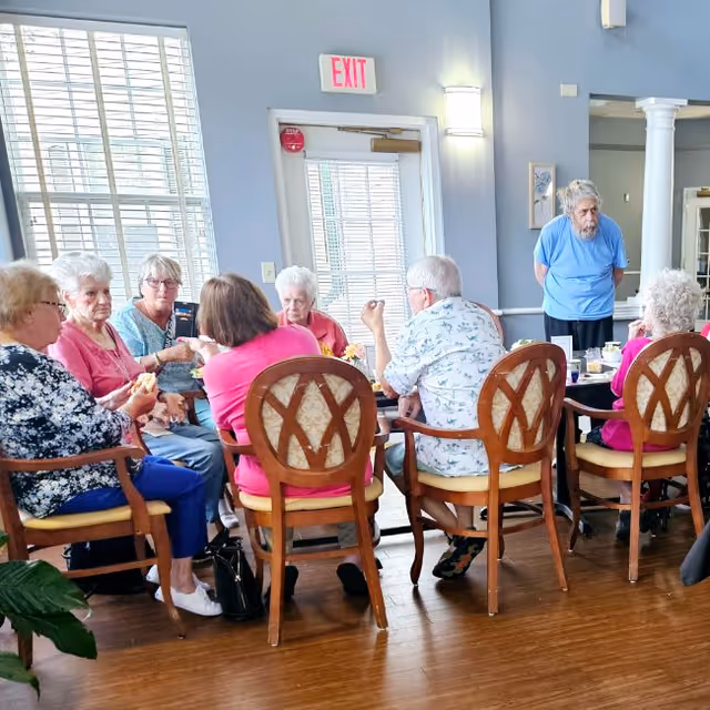 A group of elderly residents sit around a dining table in a bright common room while one person stands nearby.