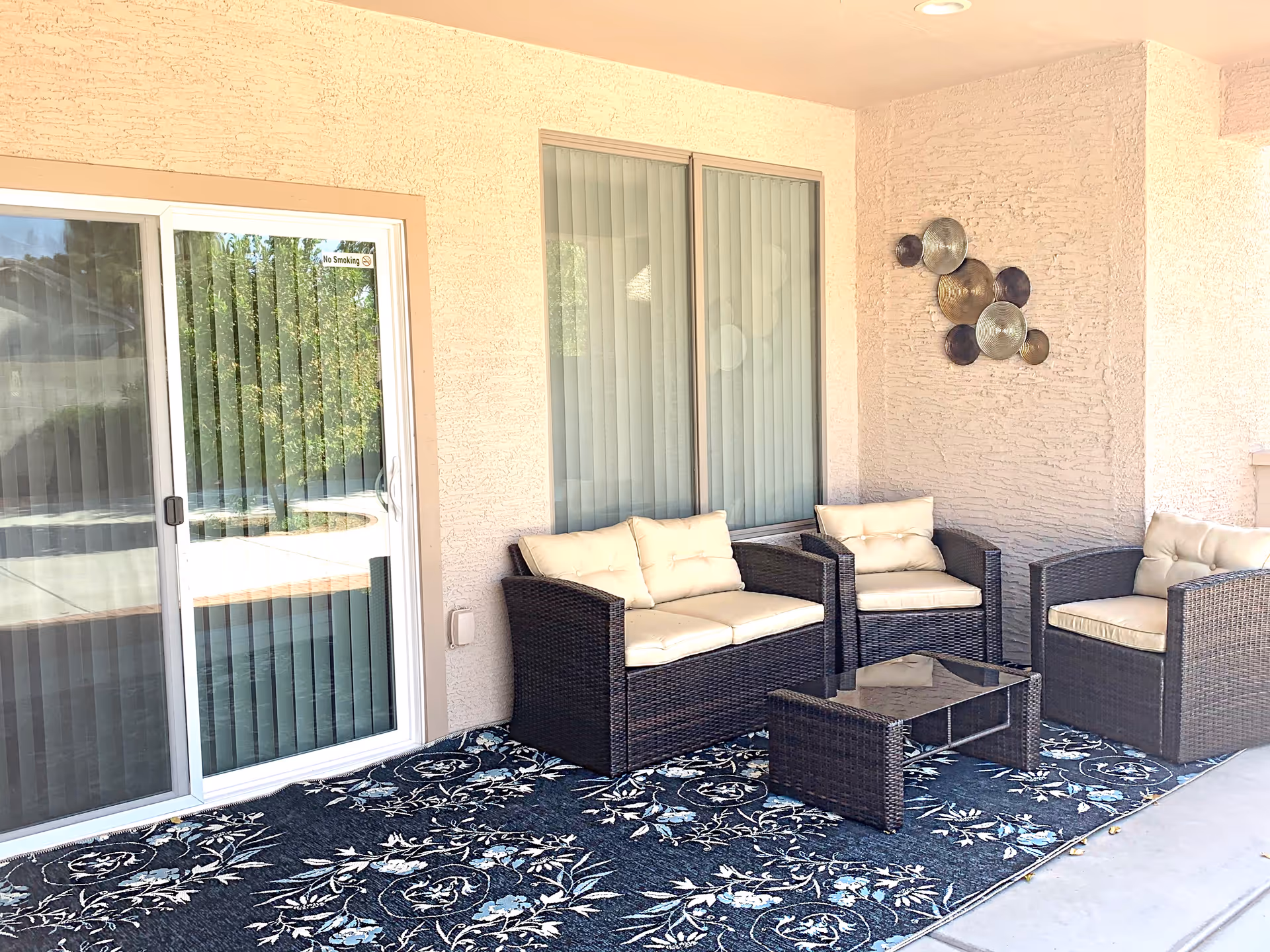 Outdoor patio area with wicker furniture including a loveseat and two armchairs with beige cushions, a glass-top coffee table, a decorative wall art piece with circular metal discs, and a sliding glass door with vertical blinds. The floor is covered with a dark floral patterned outdoor rug.