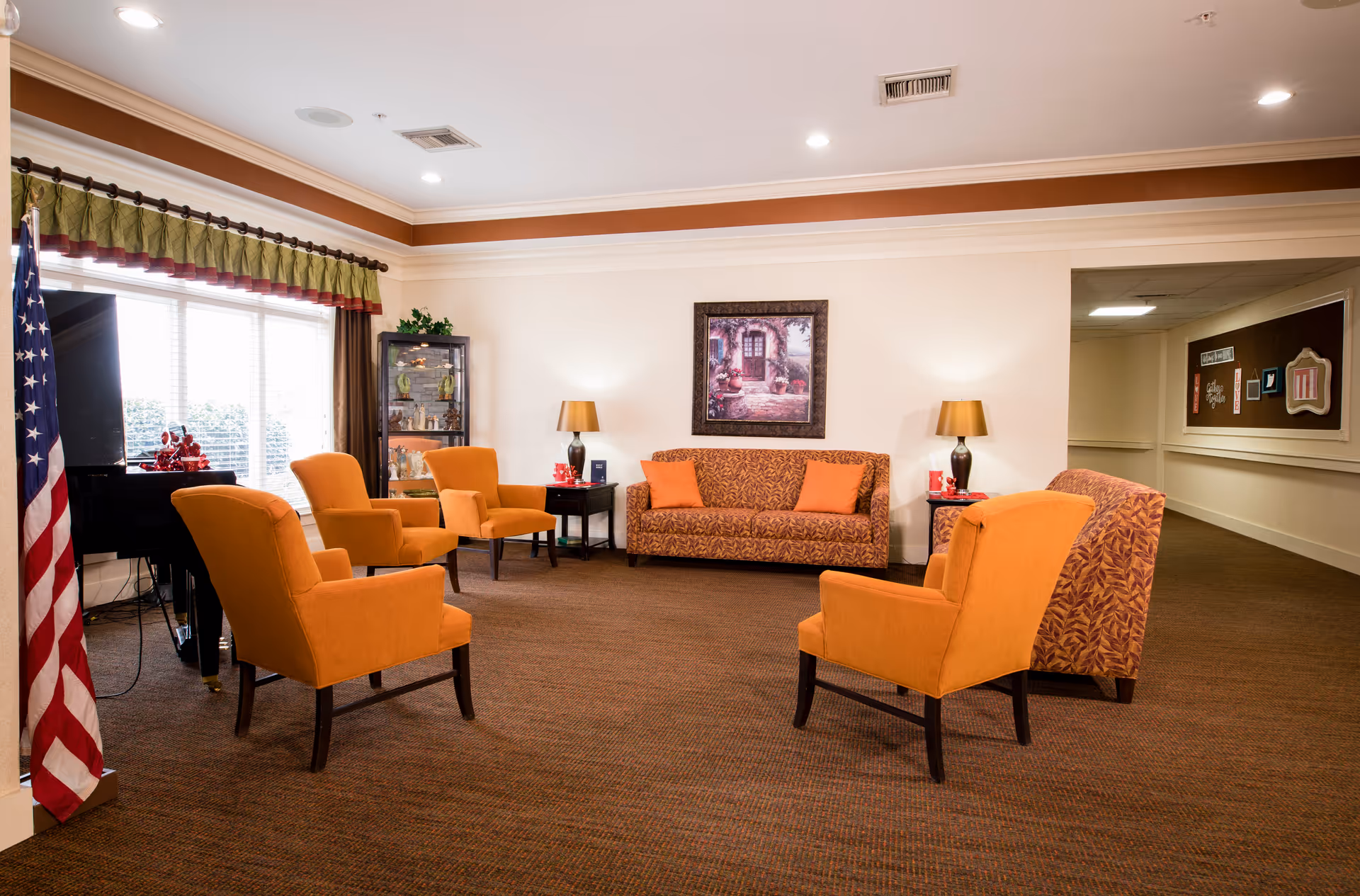 A cozy living room area in an assisted living facility featuring a patterned sofa with orange cushions, four orange armchairs arranged in a circle, two side tables with lamps, a framed painting on the wall, a glass display cabinet, a piano near a window with curtains, and an American flag in the corner.