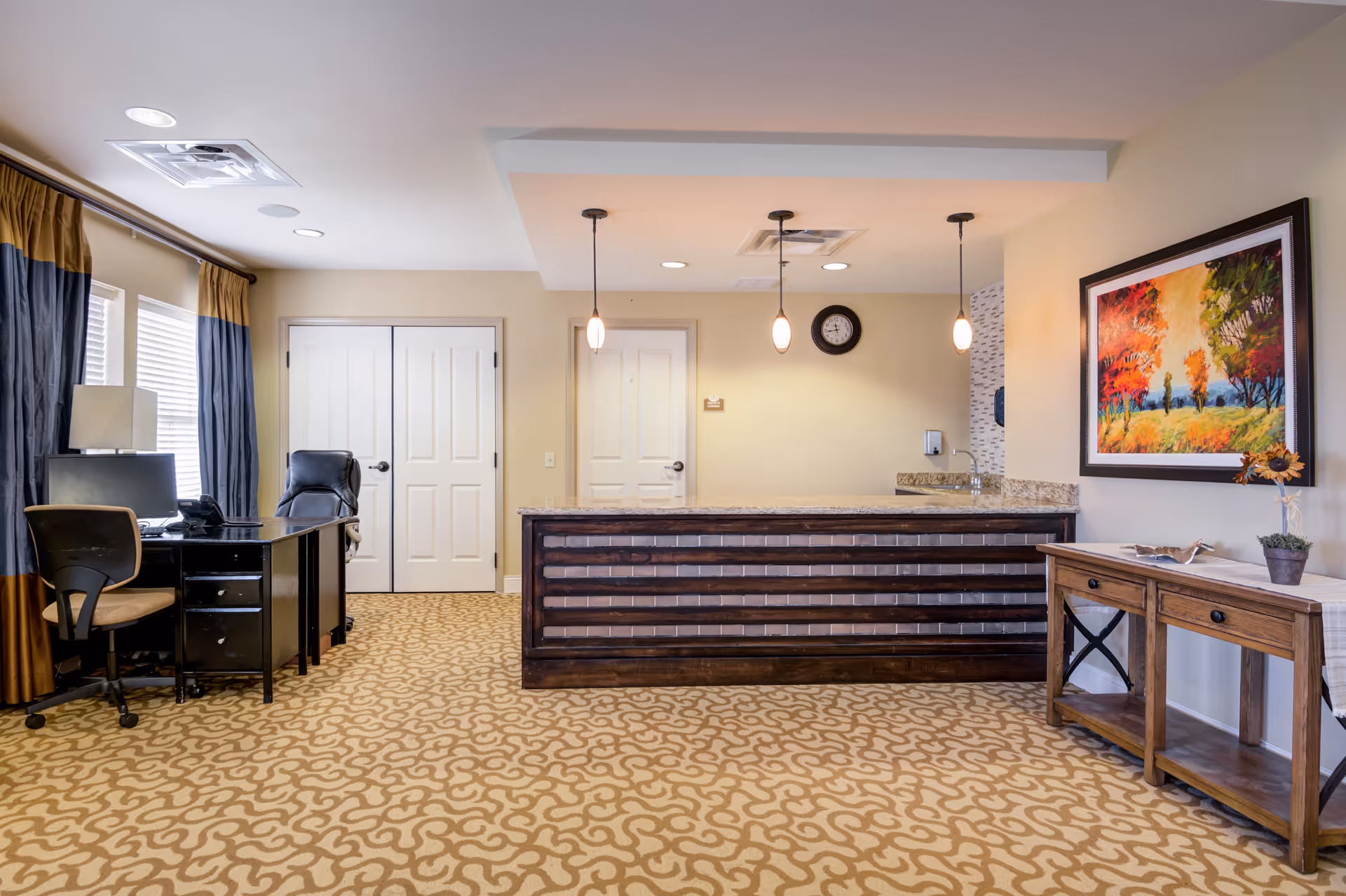Reception area with a dark wooden front desk under three hanging pendant lights, a patterned carpet, a desk with two office chairs and a computer near windows with curtains, a wooden side table with a small plant and decorative items, and a colorful framed painting on the wall.