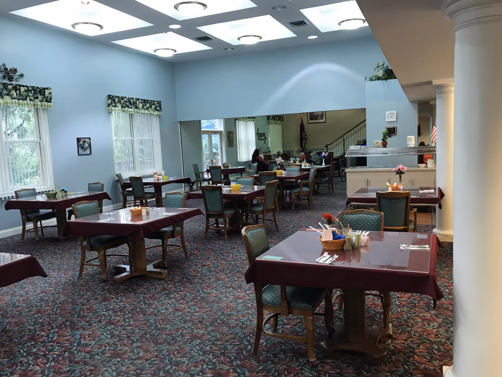 A dining room with multiple tables covered with maroon tablecloths and green cushioned chairs. The room has light blue walls, large windows with floral valances, and a patterned carpet. There are ceiling lights and skylights providing illumination. In the background, a few people are seated near a buffet area with plates and utensils.