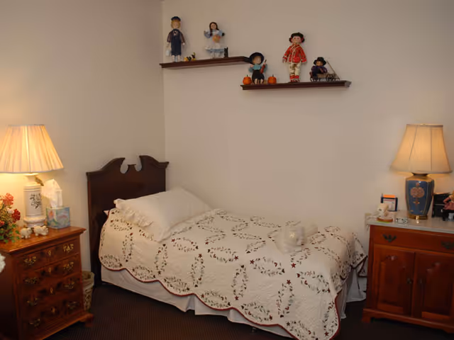 A small bedroom with a single bed covered in a white quilt with a floral pattern. There are two wooden nightstands on either side of the bed, each with a lamp. The left nightstand also has a tissue box and some decorative items. Two wooden shelves mounted on the wall above the bed display several dolls and small pumpkins. The room has a cozy and simple decor.