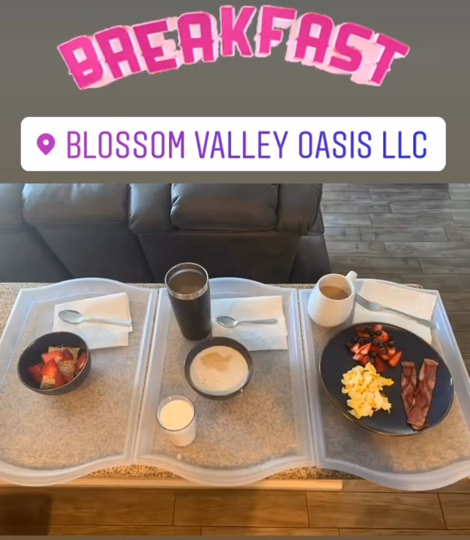 Three breakfast trays placed on a table in front of a dark-colored couch. The trays contain various breakfast items including a bowl of strawberries, a bowl of cereal with milk, a glass of milk, a cup of coffee, scrambled eggs, bacon, and mixed berries. Each tray has a napkin and a spoon or fork.