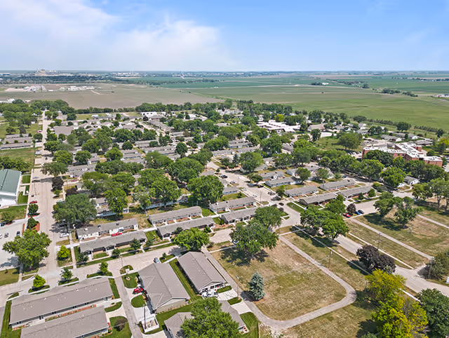 Aerial view of Good Samaritan Society - Hastings Village showing multiple single-story residential buildings surrounded by trees and green lawns, with roads and pathways connecting the community. The landscape extends to open fields in the background under a partly cloudy sky.