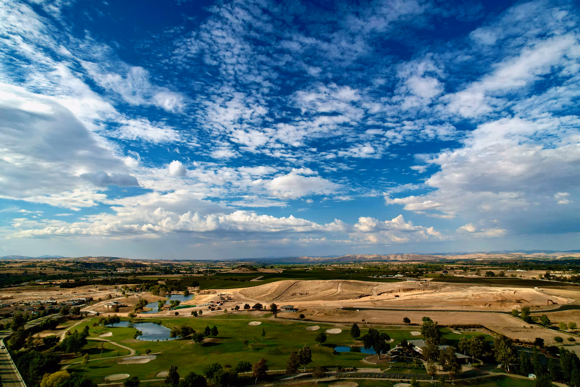 Aerial view of a golf course and surrounding fields under a vast blue sky filled with scattered clouds.