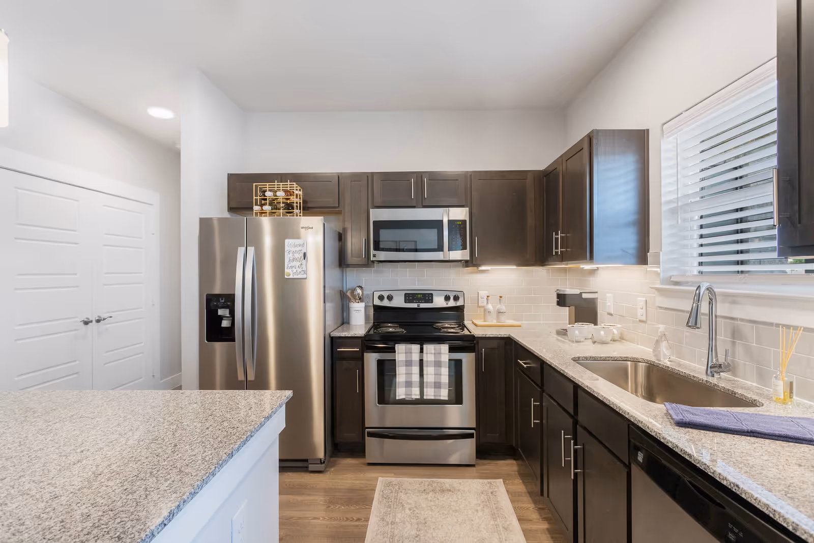 Modern kitchen with stainless steel refrigerator, oven, microwave, and dishwasher. Dark wood cabinets with silver handles, granite countertops, a sink under a window with blinds, and a kitchen island with a granite surface. A wine rack is placed on top of the refrigerator, and kitchen towels hang on the oven handle.