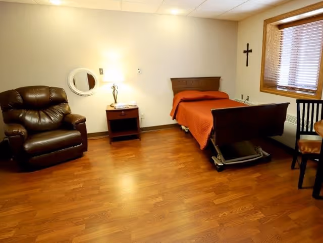 A senior living bedroom with a single bed covered by an orange blanket, a leather recliner, a nightstand with a lamp, and wood flooring.