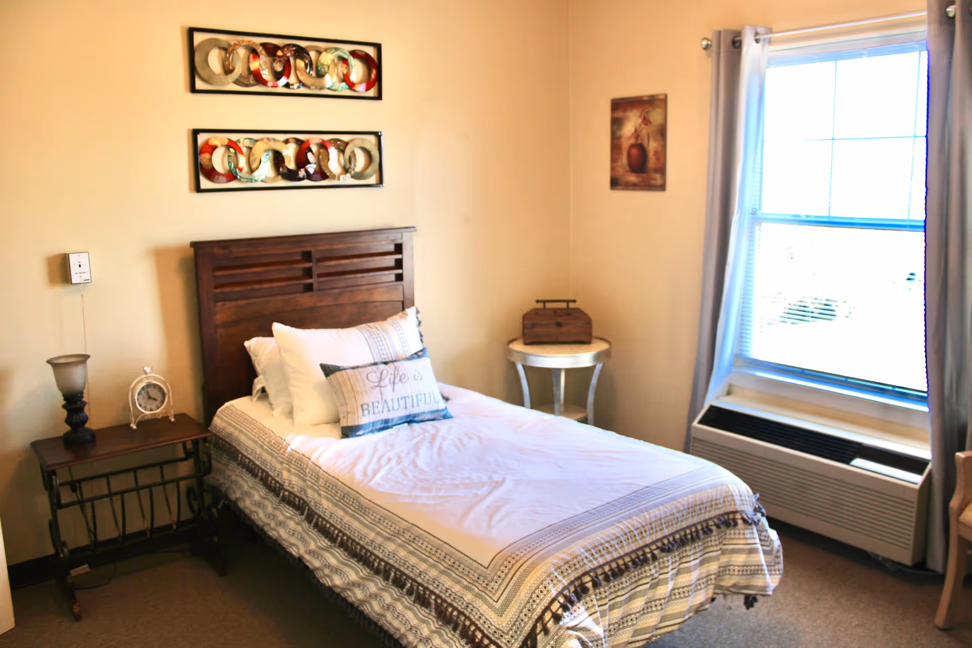 A neatly made single bed with a wooden headboard in a bedroom. The bed has white and patterned bedding with a decorative pillow that says 'Life is Beautiful'. There is a small dark wooden side table with a lamp and a clock on the left side of the bed, and a round white side table with a wooden box on the right side. A window with white curtains and blinds is on the right wall, allowing natural light into the room. Two colorful abstract wall art pieces hang above the bed, and a framed picture is on the adjacent wall.