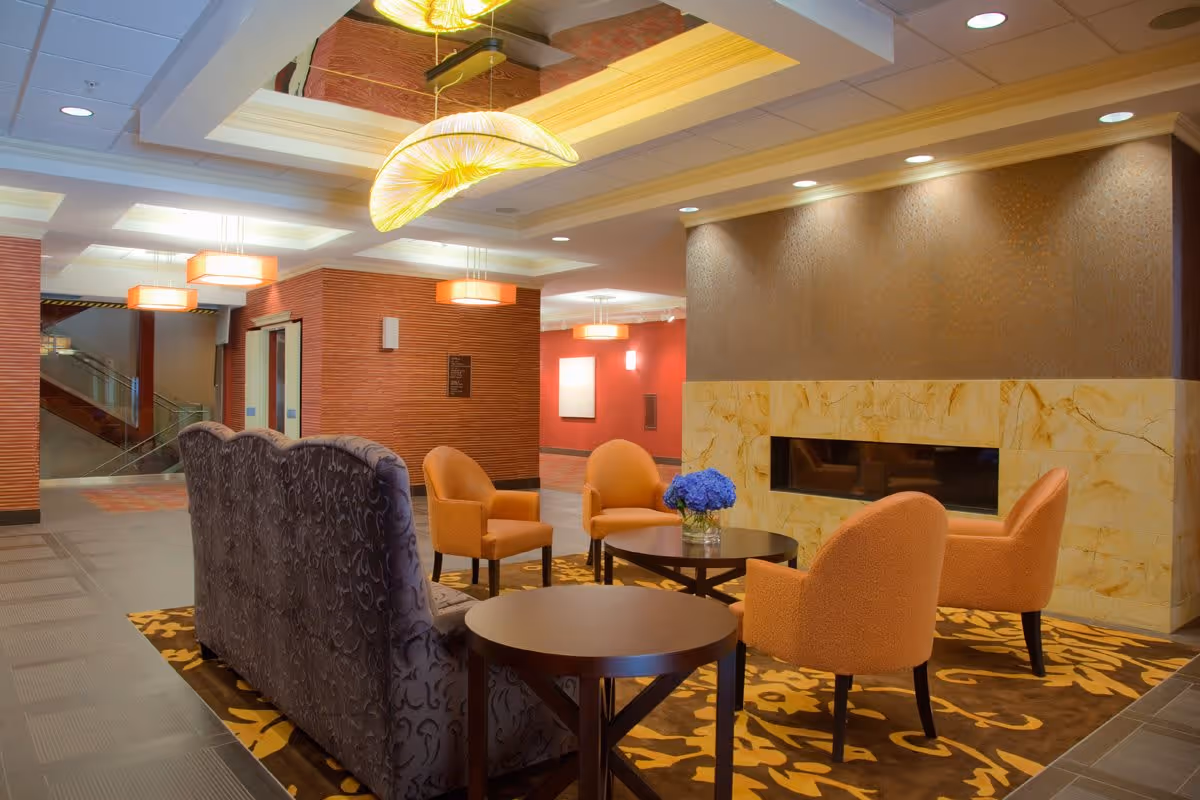 A cozy seating area in a senior living facility with four orange armchairs and a patterned dark sofa arranged around two round wooden tables. A vase with blue flowers is on one table. The area features a modern fireplace with a marble surround and warm lighting from ceiling fixtures. The walls are decorated in warm tones with textured finishes.