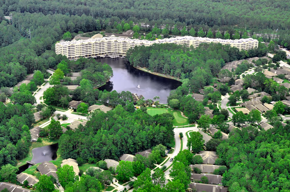 Aerial view of Cypress Village senior living facility surrounded by dense green trees, featuring a large multi-story building near a central pond with a fountain, and multiple smaller residential buildings spread out along winding roads.