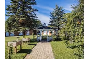 A paved walkway leading to a white gazebo surrounded by green grass, trees, and raised garden beds under a clear blue sky.