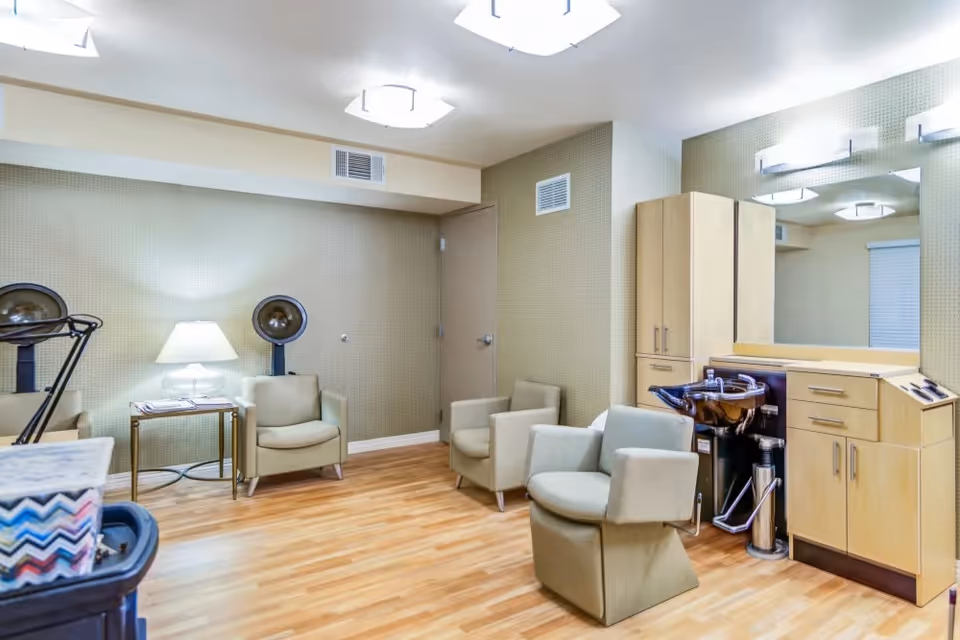 A bright salon-style room with styling chairs, hooded hair dryers, a shampoo sink, cabinets and a large mirror.