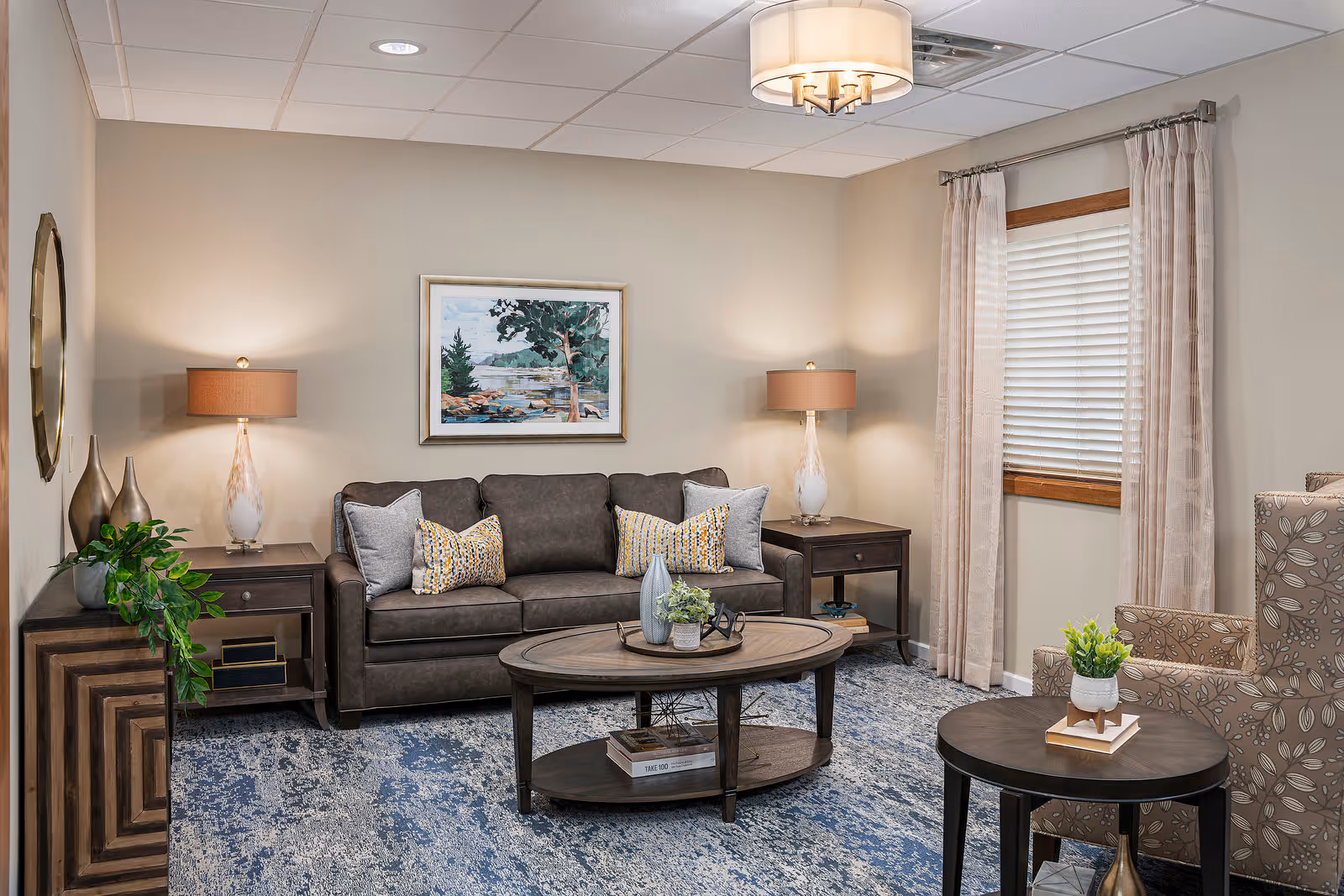 A cozy living room with a dark brown leather sofa adorned with four decorative pillows, two side tables each with a lamp, a round wooden coffee table with decorative items, a patterned armchair, a small round side table with a plant, a window with beige curtains, and a framed landscape painting on the wall.
