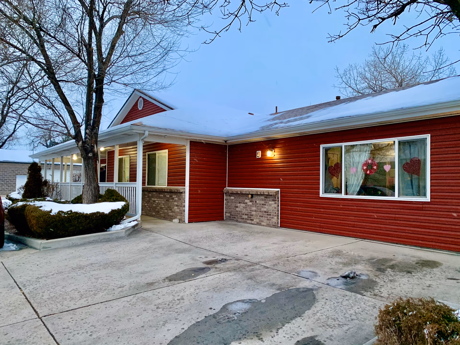 Exterior view of a single-story building with red siding and a white roof covered in snow. There is a concrete driveway in front, a tree with no leaves, and some bushes partially covered with snow. The building has a porch with white railings and a window decorated with heart-shaped ornaments.