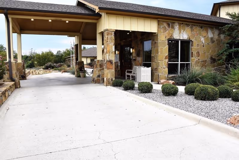 Exterior view of a building with stone walls and a covered driveway entrance. There are neatly trimmed bushes and gravel landscaping along the side of the driveway, with a bench and some plants near the entrance.