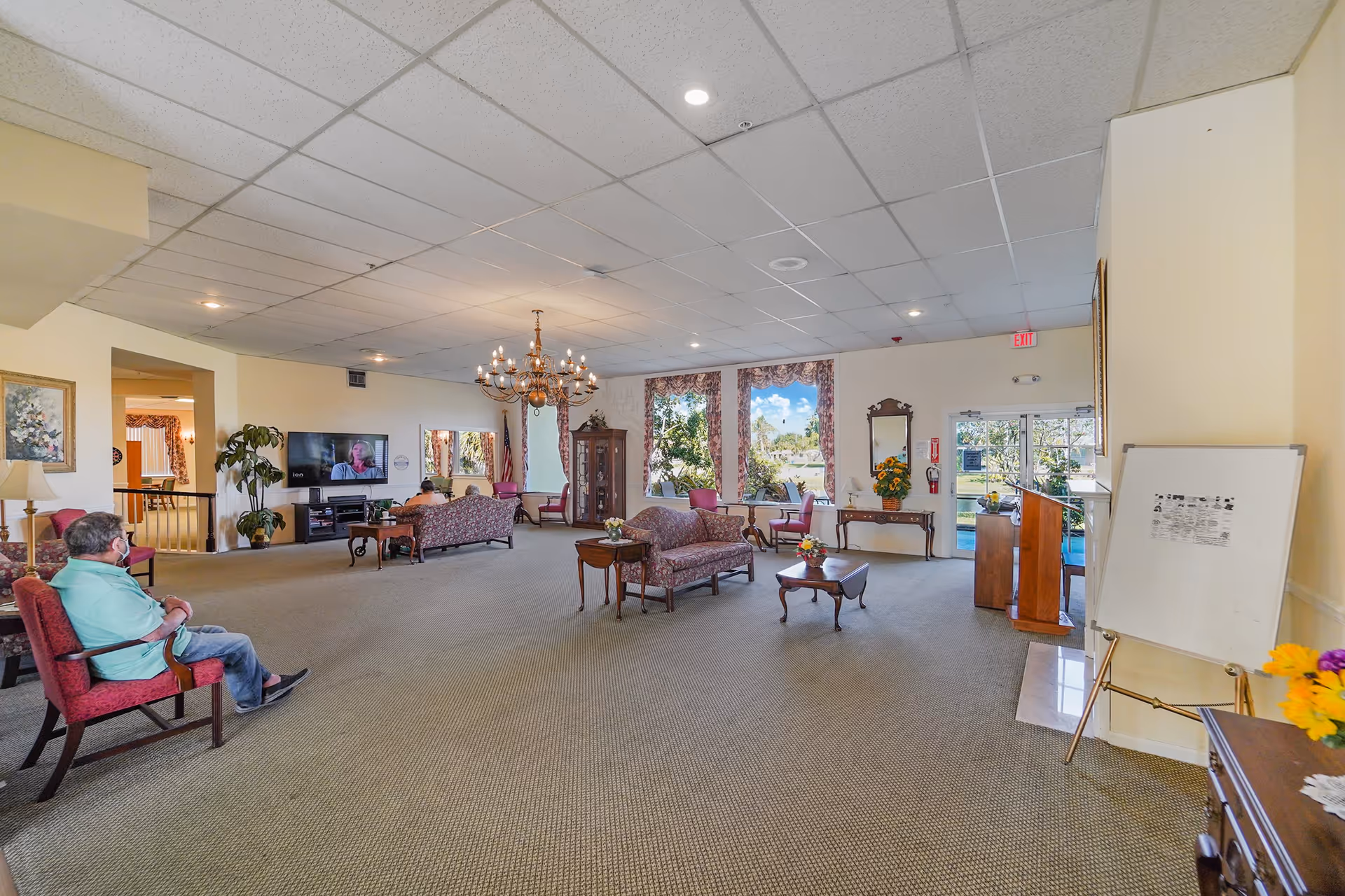 Spacious assisted-living common room with sofas, chairs, a chandelier, television, and a person seated on the left.