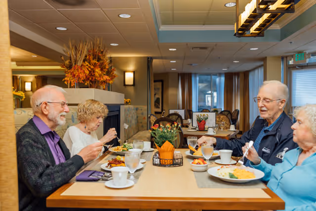 Four elderly people sitting around a dining table in a well-lit room, eating a meal together. The table is set with plates of food, glasses of water, coffee cups, and a small decorative plant in the center. The room has warm lighting, autumn-themed decorations, and large windows with curtains in the background.