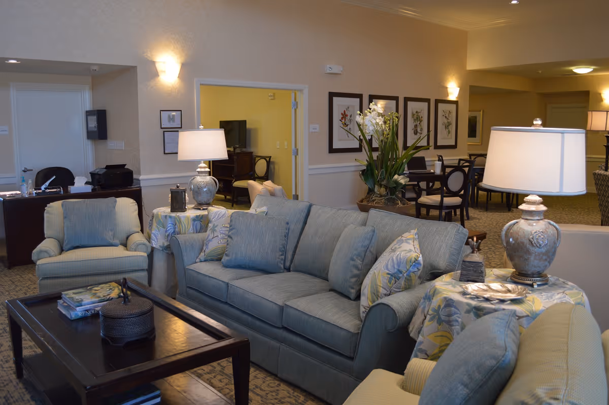 A cozy living room area in a senior living facility with a blue sofa, two armchairs, a dark wooden coffee table with books and decorative items, two side tables with lamps, and a dining area with tables and chairs in the background. The walls are decorated with framed botanical prints and there is a large potted plant near the dining area.