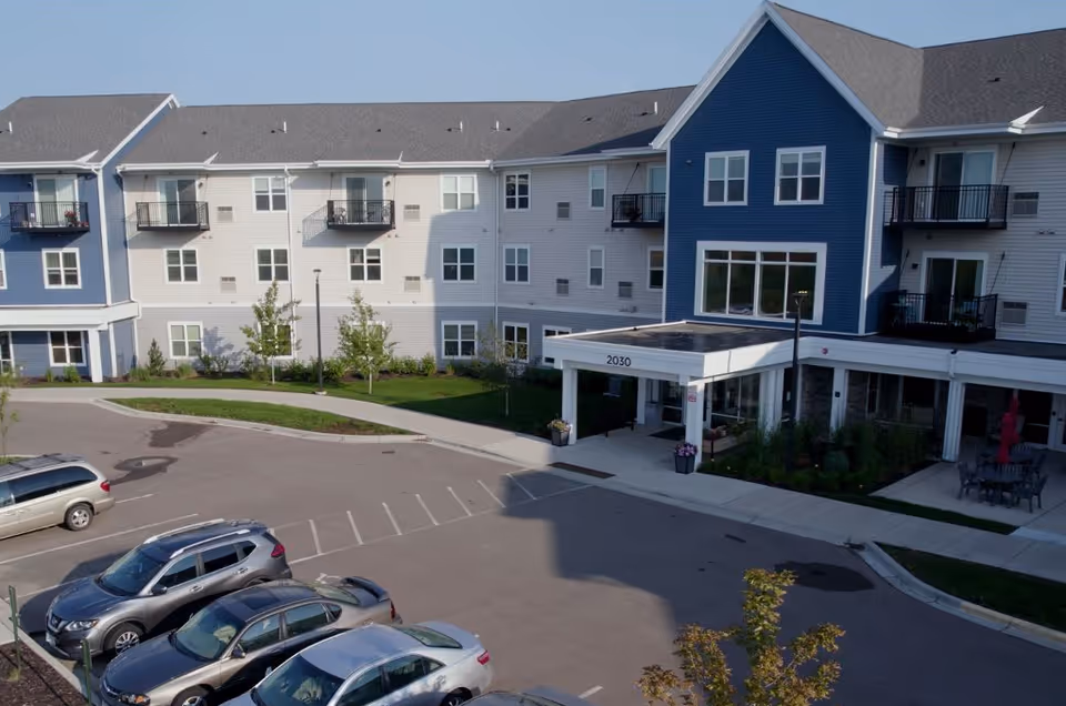 Exterior view of a multi-story senior living facility building with a blue and white facade, balconies, and a covered entrance labeled 2030. Several cars are parked in the parking lot in front of the building, and there are small trees and landscaped areas around the entrance.
